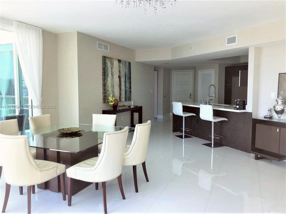 Open-concept living space featuring high-gloss tile flooring, crystal chandelier, kitchen with breakfast bar, dark-finish cabinetry, and floor-to-ceiling glass sliding doors