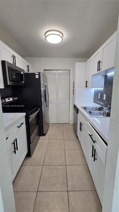 Kitchen featuring white shaker-style cabinetry with matte black hardware, light-toned solid surface countertops, stainless steel appliances, tiled backsplash, and ceramic floor tiling