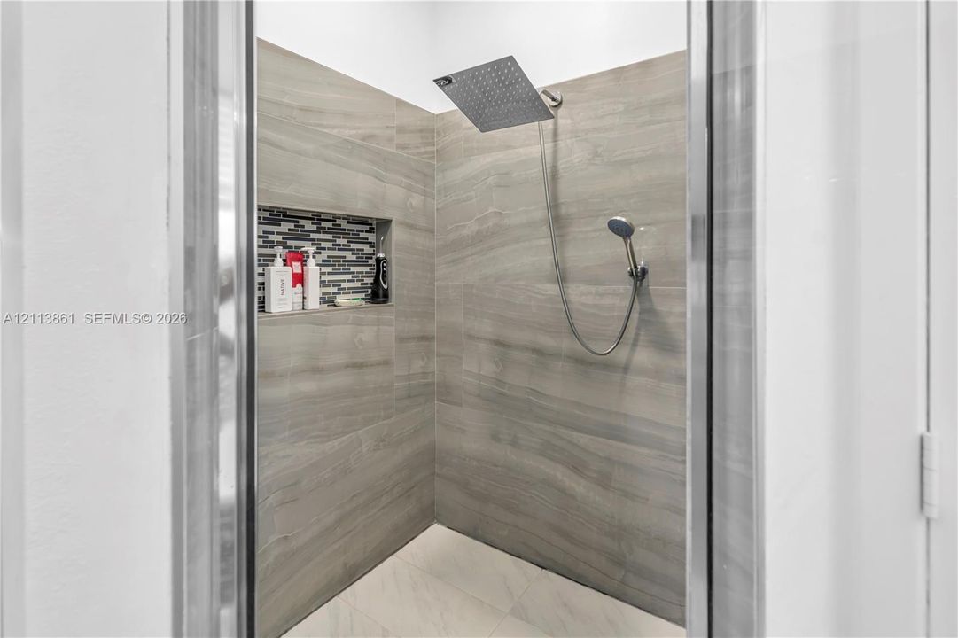 Walk-in shower featuring large-format neutral tile wall cladding, a built-in recessed niche with mosaic accent tiling, a rainfall showerhead, and a handheld shower wand