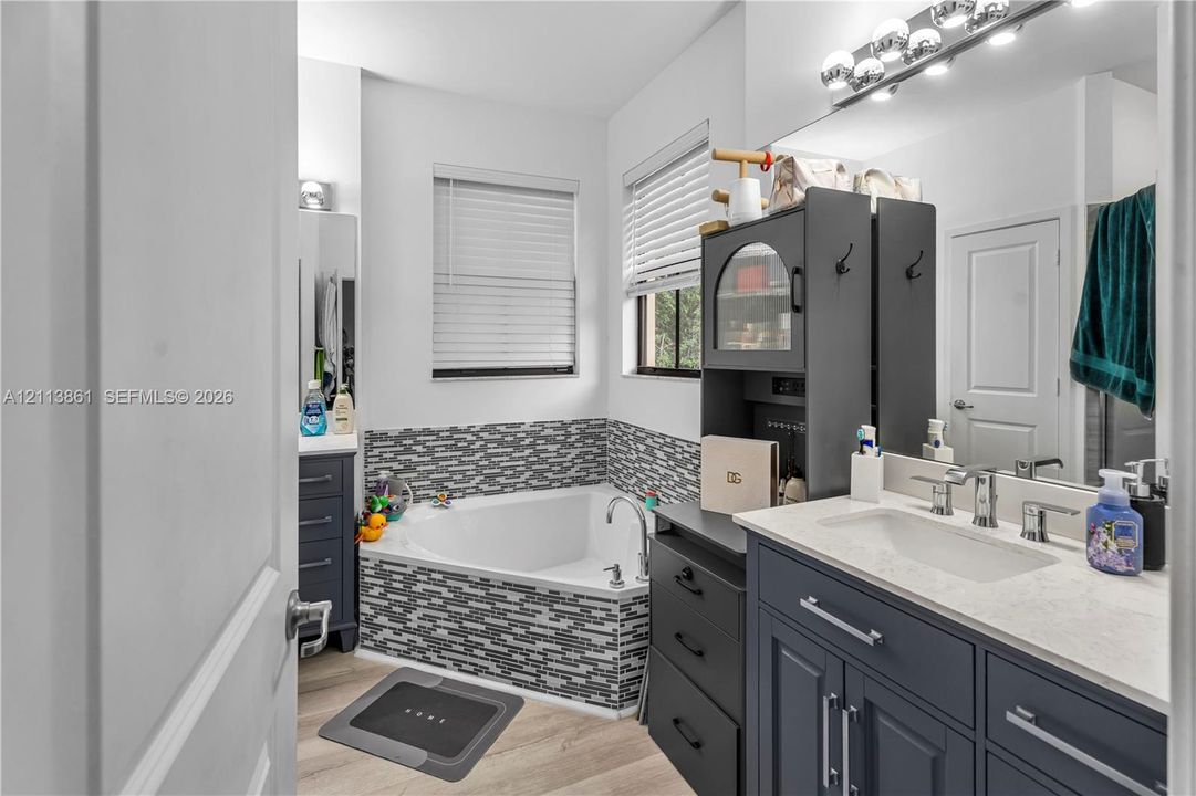 Bathroom featuring a vanity with a light-toned countertop, undermount sink, and dark cabinetry, complemented by a corner soaking tub with mosaic tile surround and wood-finish flooring