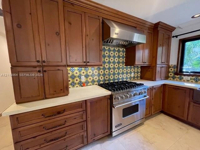 Custom wood cabinetry, stainless steel professional-grade gas range, stainless steel vent hood, patterned tile backsplash, and light-toned solid surface countertops