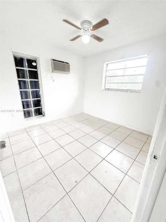 Interior room featuring large-format ceramic tile flooring, a multi-blade ceiling fan with integrated lighting, a vertical rectangular window, a horizontal sliding window, and a wall-mounted air conditioning unit