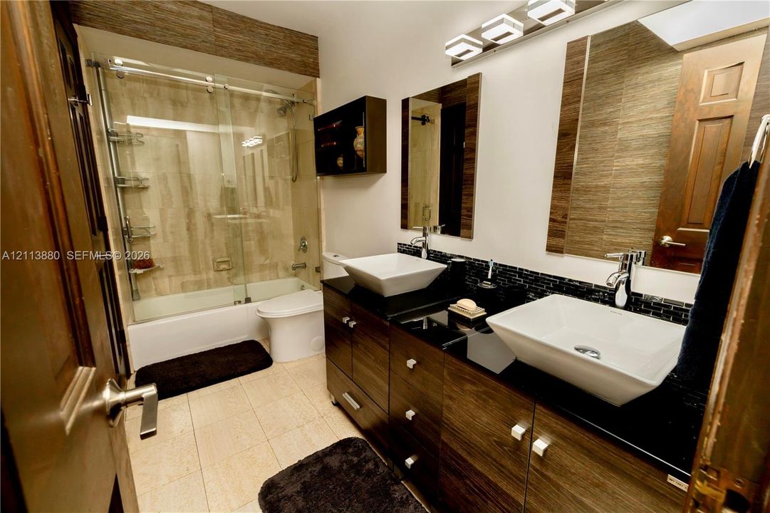 Dual vessel sinks with polished chrome faucets, black countertop, wood-finish cabinetry, tile-surround shower with sliding glass door, and integrated vanity lighting
