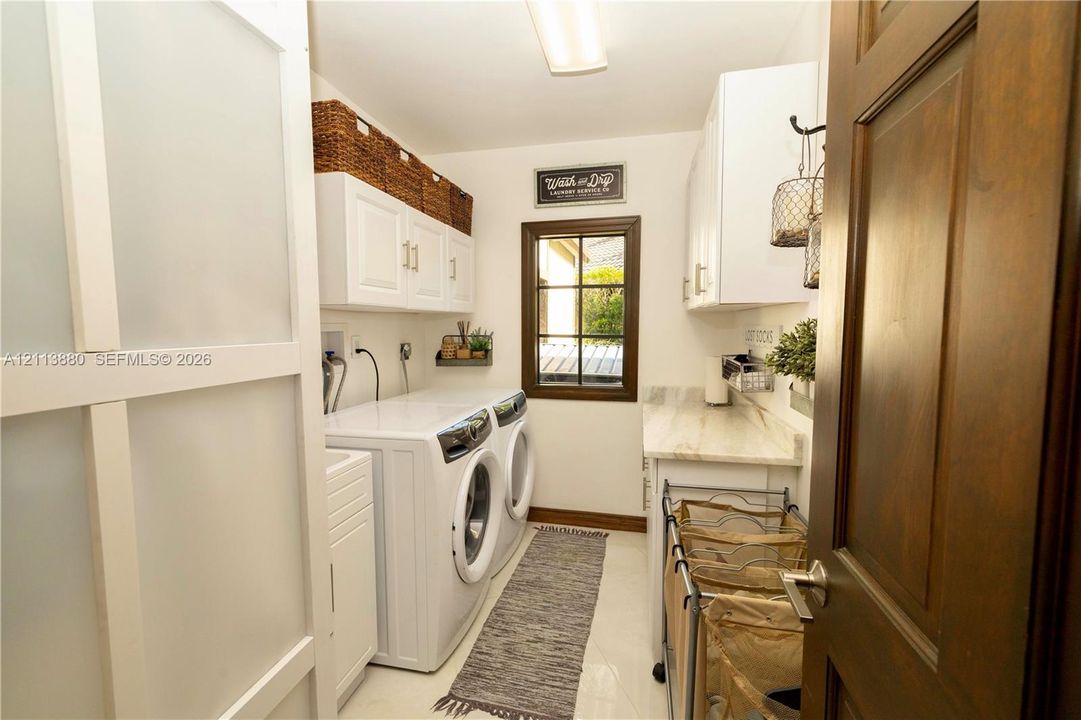 Laundry area featuring white shaker-style cabinetry, stone-surface countertop, dual-pane window with dark trim, and a sliding frosted glass door