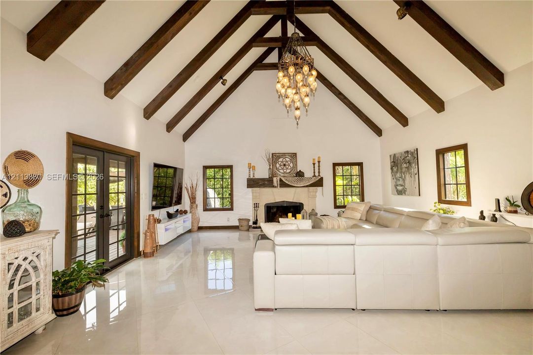 Vaulted ceiling with exposed wood-finish beams, polished tile flooring, stone-surround fireplace with a wooden mantel, and multi-pane glass french doors