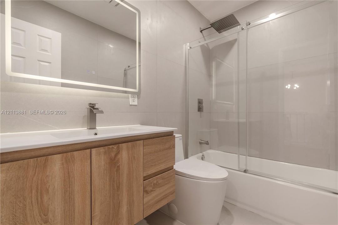 Bathroom featuring a wood-finish vanity with a rectangular sink, wall-mounted rectangular LED-lit mirror, and a tub with a glass sliding door enclosure and rain-style shower head