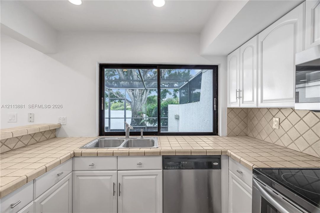 Kitchen featuring white cabinetry, tile countertops, diamond-patterned tile backsplash, stainless steel dishwasher, and sliding glass window