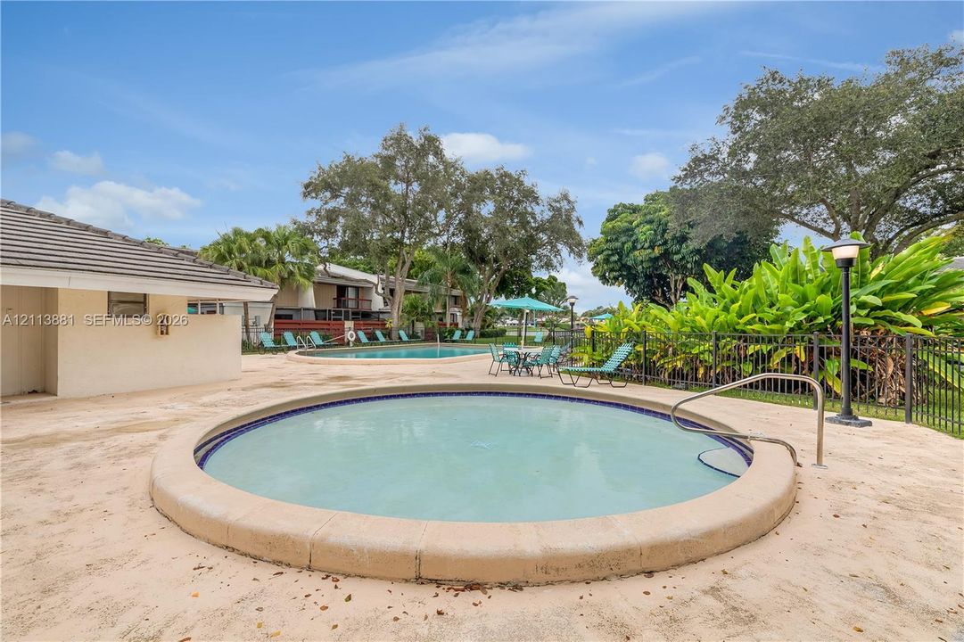 Circular spa featuring a concrete surround, metal handrail, and tile inlay, complemented by a secondary swimming pool, expansive concrete decking, and mature landscaping