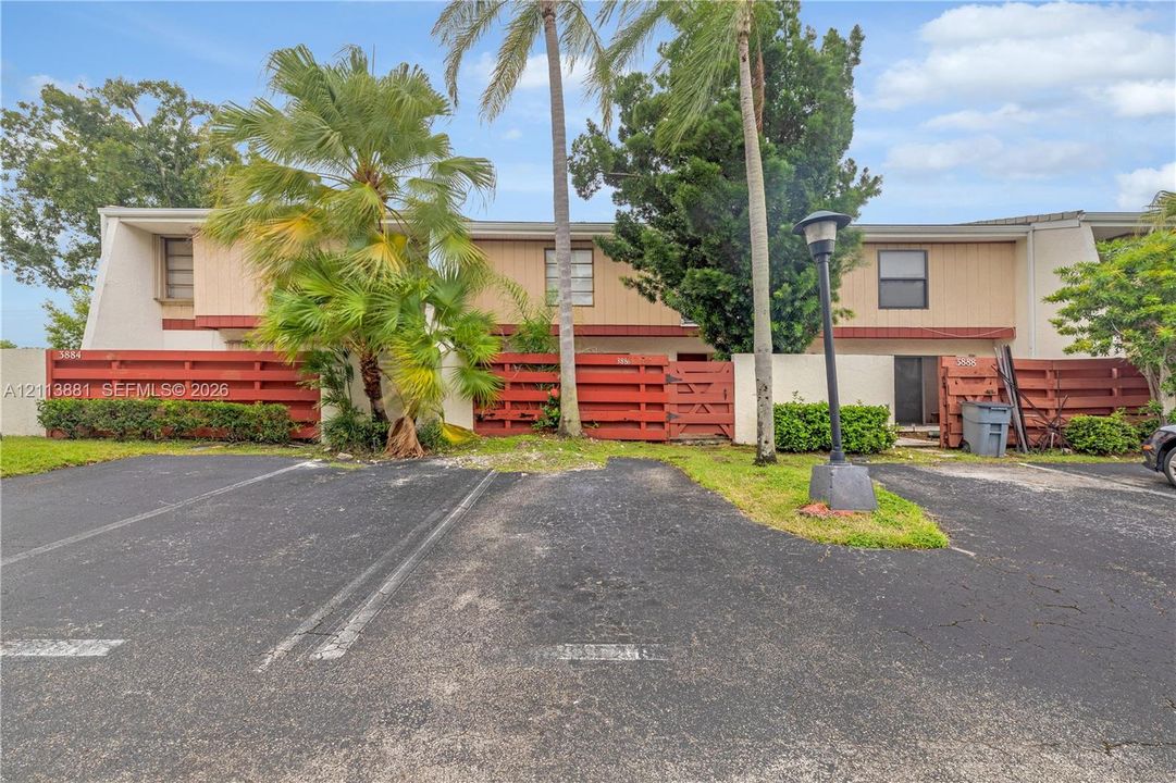 Two-story exterior featuring wood-paneled siding, decorative red wood fencing, asphalt parking area, and mature palm landscaping