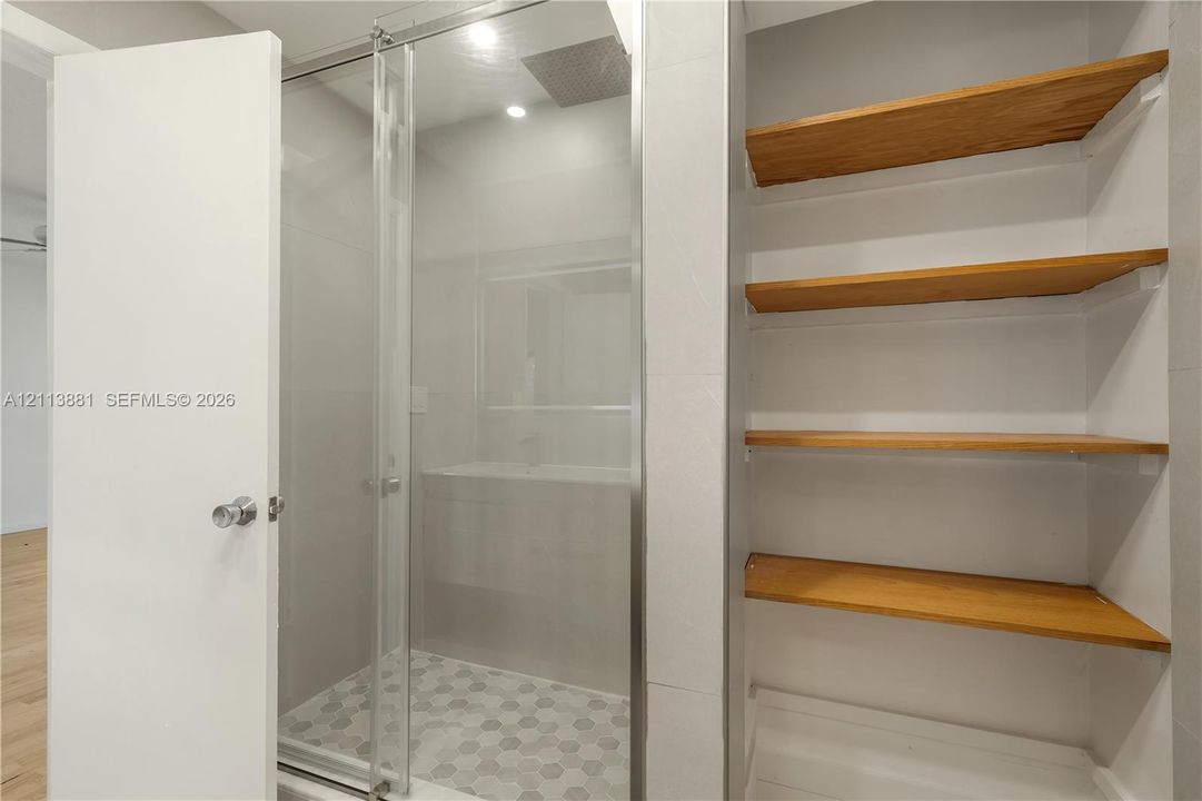 Walk-in shower featuring a glass enclosure, oversized rain shower head, hexagonal tile flooring, and integrated wood-finish shelving