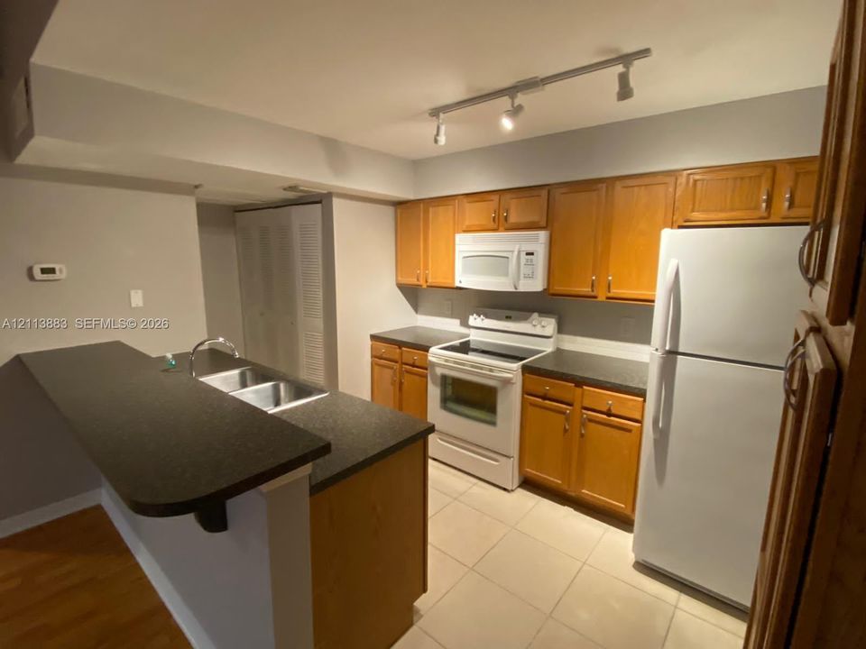 Kitchen featuring wood cabinetry, dark laminate countertops, a dual-basin sink, white appliances, and track lighting
