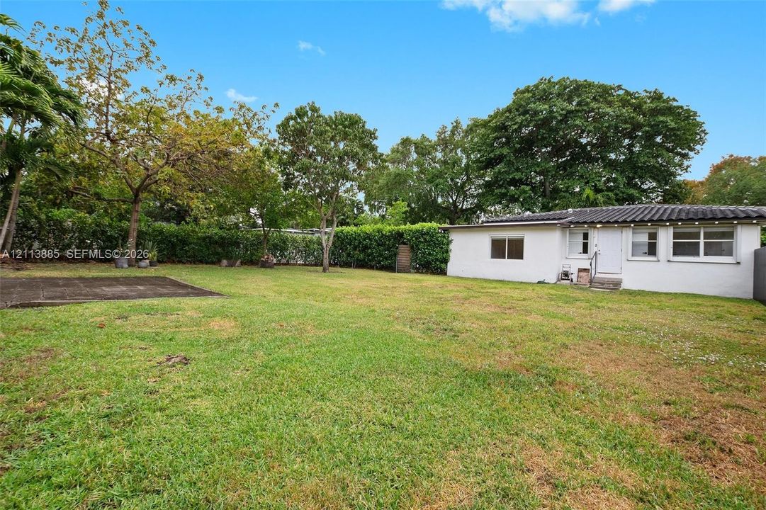 Expansive backyard featuring a large grass lawn, perimeter privacy hedges, mature shade trees, a paved patio area, and a white exterior structure with horizontal windows