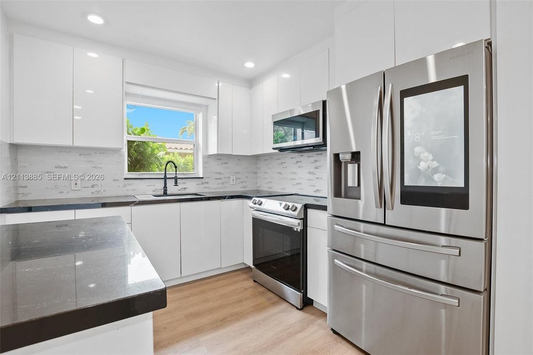 Kitchen featuring high-gloss white cabinetry, dark stone-finish countertops, stainless steel appliances, a textured tile backsplash, and wood-finish flooring