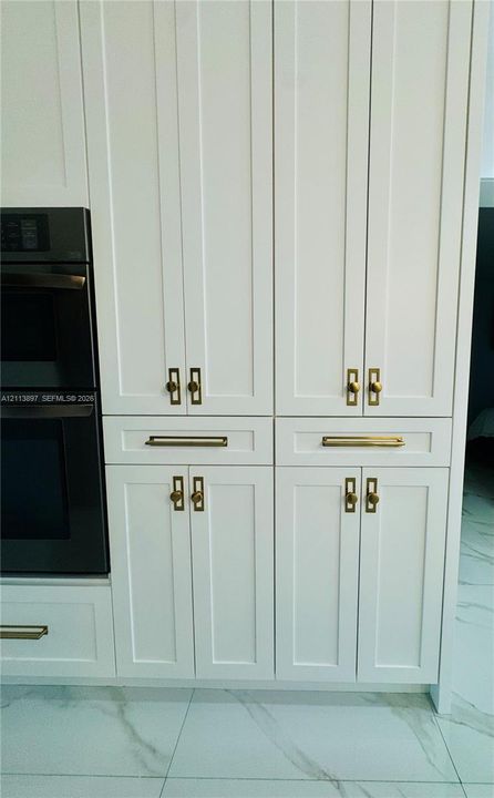 White shaker-style cabinetry with recessed panels, brushed brass hardware, and a built-in double wall oven, set against large-format rectified tile flooring