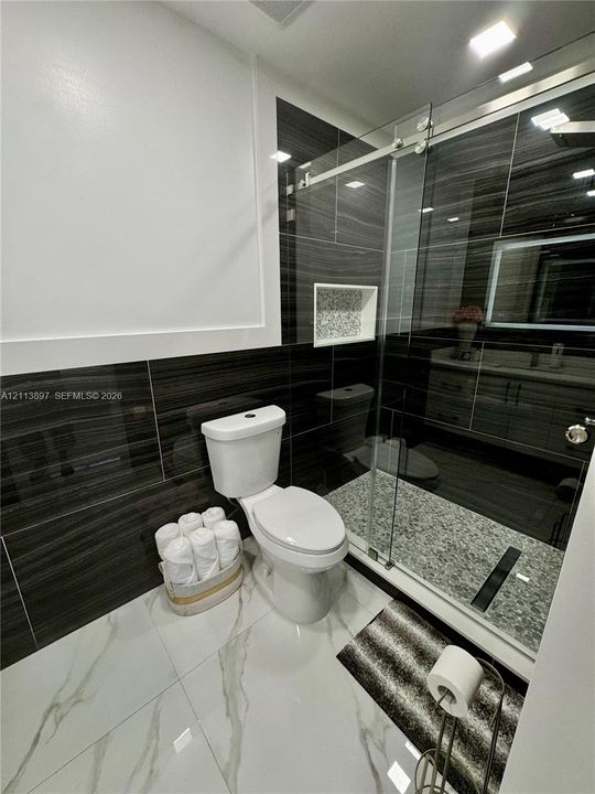Bathroom featuring large-format white tile flooring, dark horizontal-patterned wall tiling, a walk-in shower with a glass sliding door, a recessed shower niche, and a vanity cabinet with an integrated LED-lit mirror