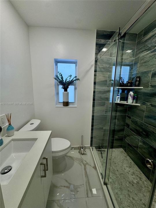 Bathroom featuring large-format stone-finish floor tiling, a vanity with a rectangular integrated sink, a walk-in shower with stone-textured wall tiles and a glass sliding door, and integrated shower shelving