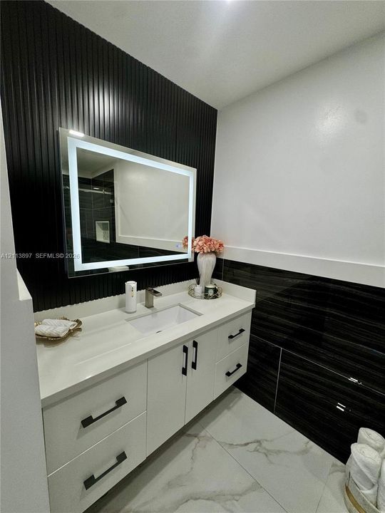 White vanity with clean-lined cabinetry, quartz countertop, integrated rectangular sink, illuminated mirror, and vertical slat accent wall