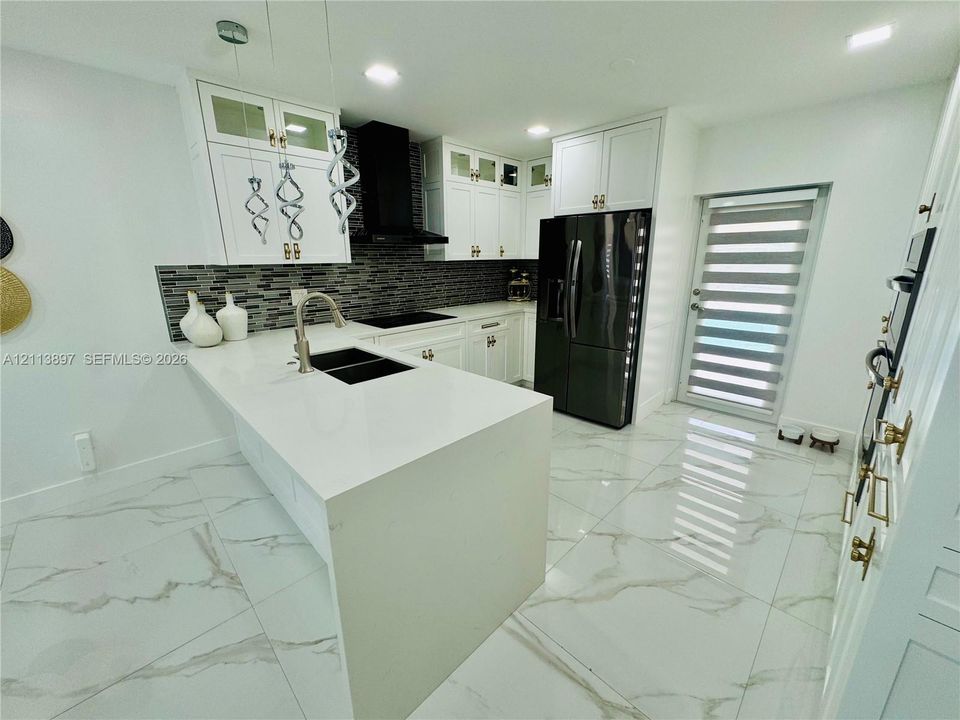 Kitchen featuring white cabinetry with glass-panel accents, stone-composite countertops, a multi-tonal linear tile backsplash, polished stone-finish flooring, and a central island with a double-basin under-mount sink