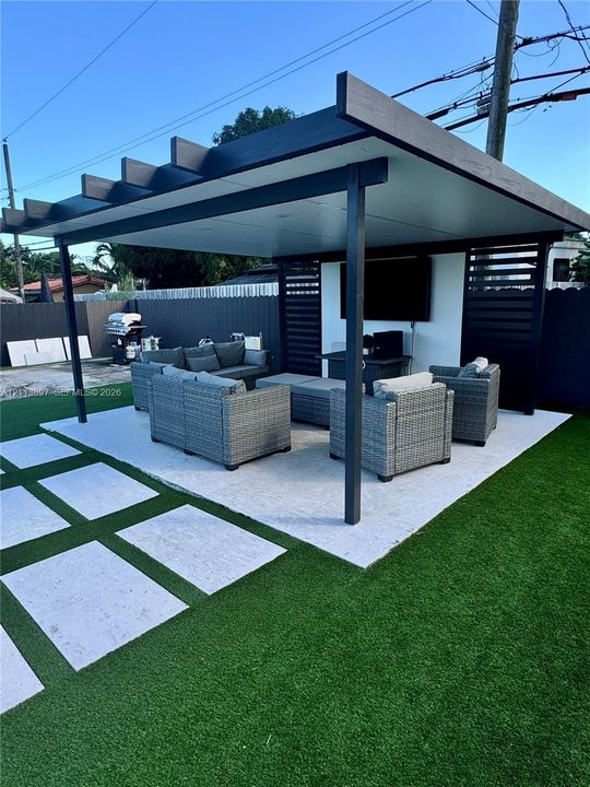Covered outdoor pavilion featuring a flat roof structure, built-in slatted privacy screening, integrated lighting, stone-tiled flooring, and surrounding artificial turf with inset paver pathway