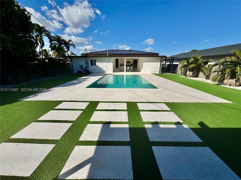 Backyard featuring an inground pool, large stone-finish deck, geometric paver pathway, and synthetic lawn