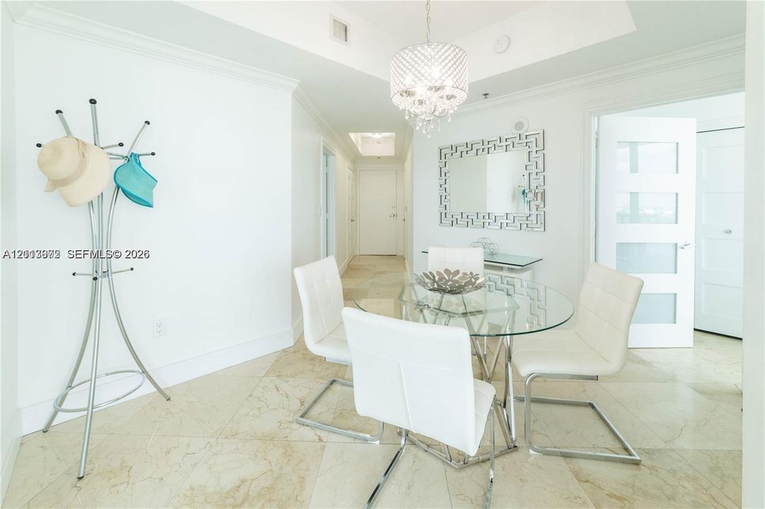 Light-toned stone tile flooring throughout, recessed tray ceiling, crystal-style chandelier, and white-framed door with translucent panels