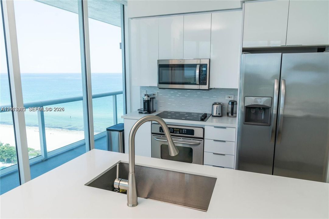 Kitchen featuring white high-gloss cabinetry, stainless steel appliances, quartz countertops, a central island with an undermount sink, and floor-to-ceiling windows