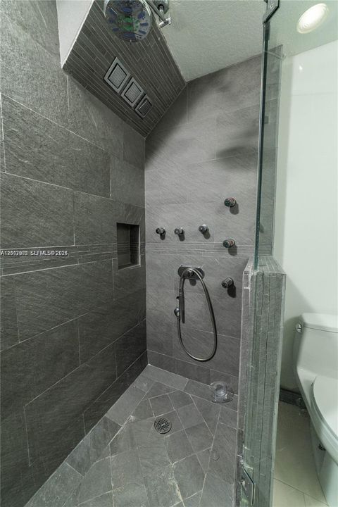 Walk-in shower featuring large-format dark grey stone tile walls, a built-in recessed niche, multiple body sprayers, a handheld shower head, and a glass partition