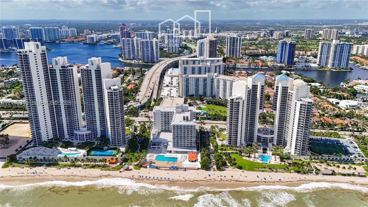 Aerial perspective of waterfront high-rise residential towers featuring direct beach access, outdoor swimming pools, and tennis courts
