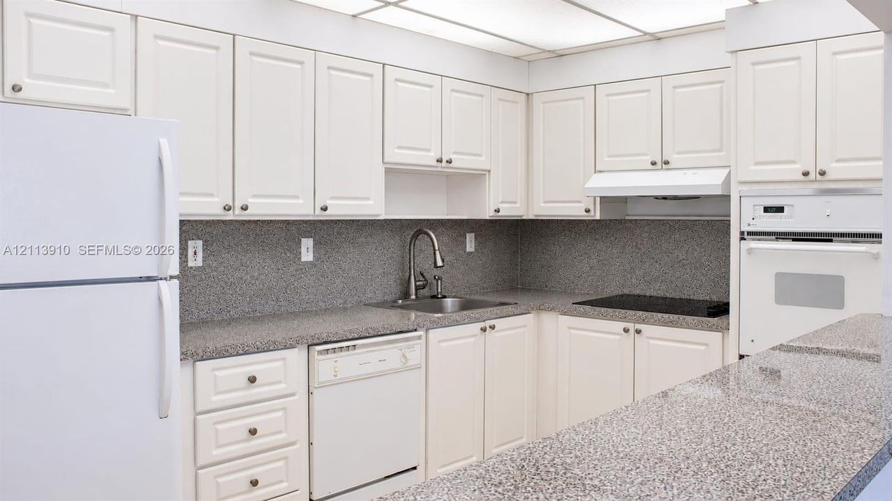 Kitchen featuring off-white cabinetry, speckled stone countertops, a stainless steel faucet, a built-in wall oven, and a drop ceiling with integrated lighting