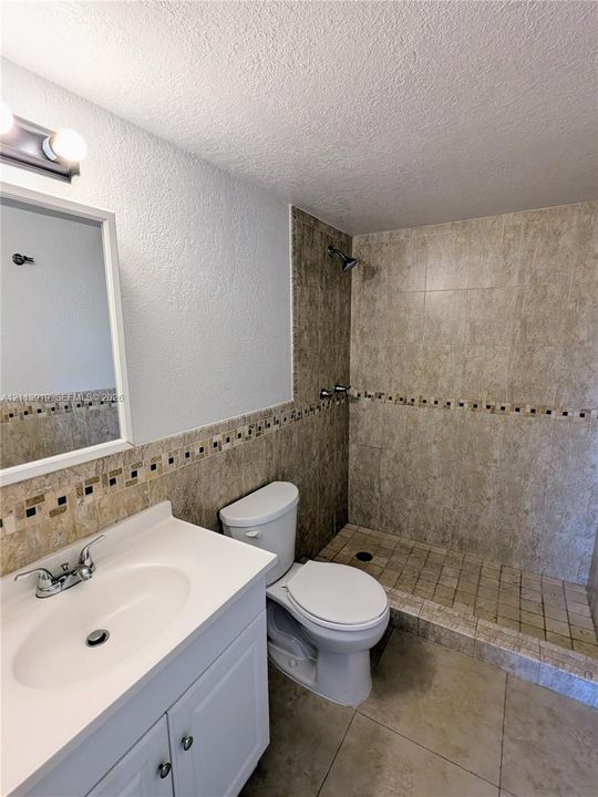Bathroom featuring a white vanity cabinet with an integrated basin, wall-mounted vanity lighting, a walk-in shower with stone-finish tile surround, and decorative accent tiling