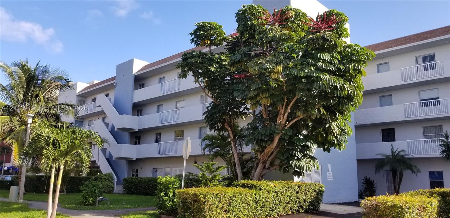 Multi-story exterior featuring white stucco siding, tiered balconies with metal railings, external staircases, and manicured landscaping with mature trees