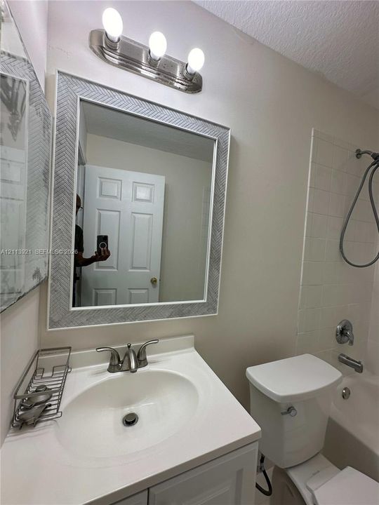 Bathroom featuring a vanity with a white countertop and oval sink, a wall-mounted mirror with a textured frame, a three-light vanity fixture, and a tub-shower combination with tiled surround