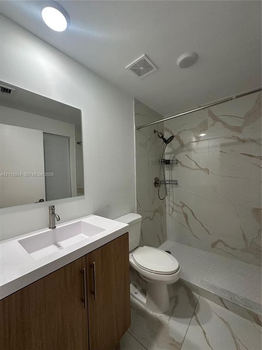Bathroom featuring a wood-finish vanity with a white countertop and integrated sink, walk-in shower with floor-to-ceiling stone-patterned tile, and large-format tile flooring
