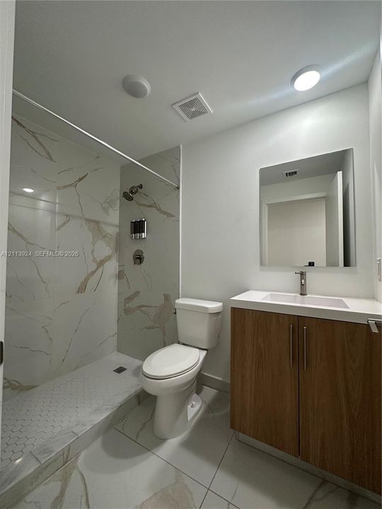 Bathroom featuring a walk-in shower with stone-patterned tile surround, wood-finish vanity with an integrated basin, frameless mirror, and large-format tile flooring