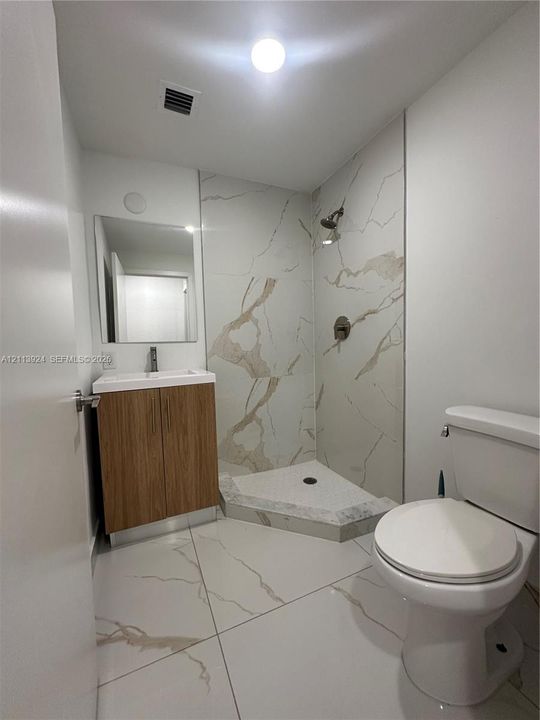 Bathroom featuring large-format veined tile flooring, a wood-finish vanity with a white basin, a corner shower enclosure with tiled surround, and a wall-mounted mirror