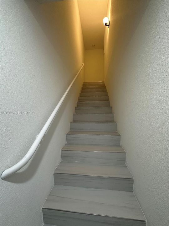 Staircase featuring wood-finish treads, a white wall-mounted handrail, textured wall surfaces, and an attached wall sconce