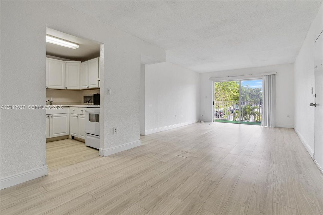 Open-concept interior featuring wood-finish flooring, sliding glass door with access to an exterior balcony, and kitchen with white cabinetry and flush-mount lighting