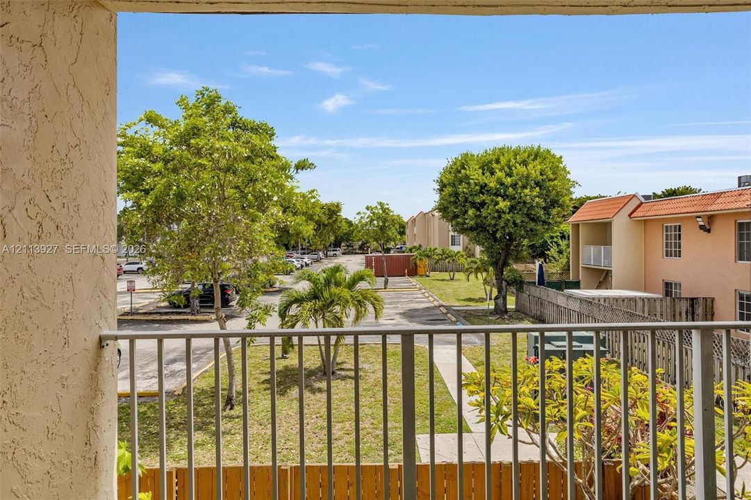 Balcony featuring metal railing and stucco pillar finish, overlooking exterior grounds with mature landscaping and paved parking area