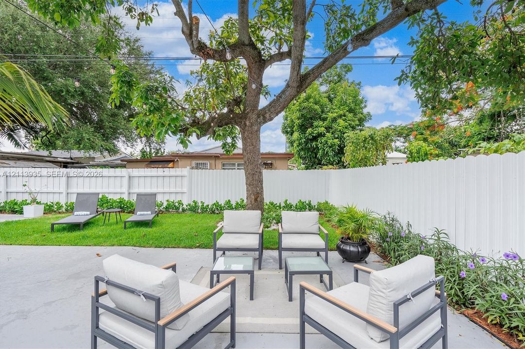 Private backyard featuring a poured concrete patio, manicured lawn, perimeter white vinyl fencing, mature canopy tree, and landscaped garden beds