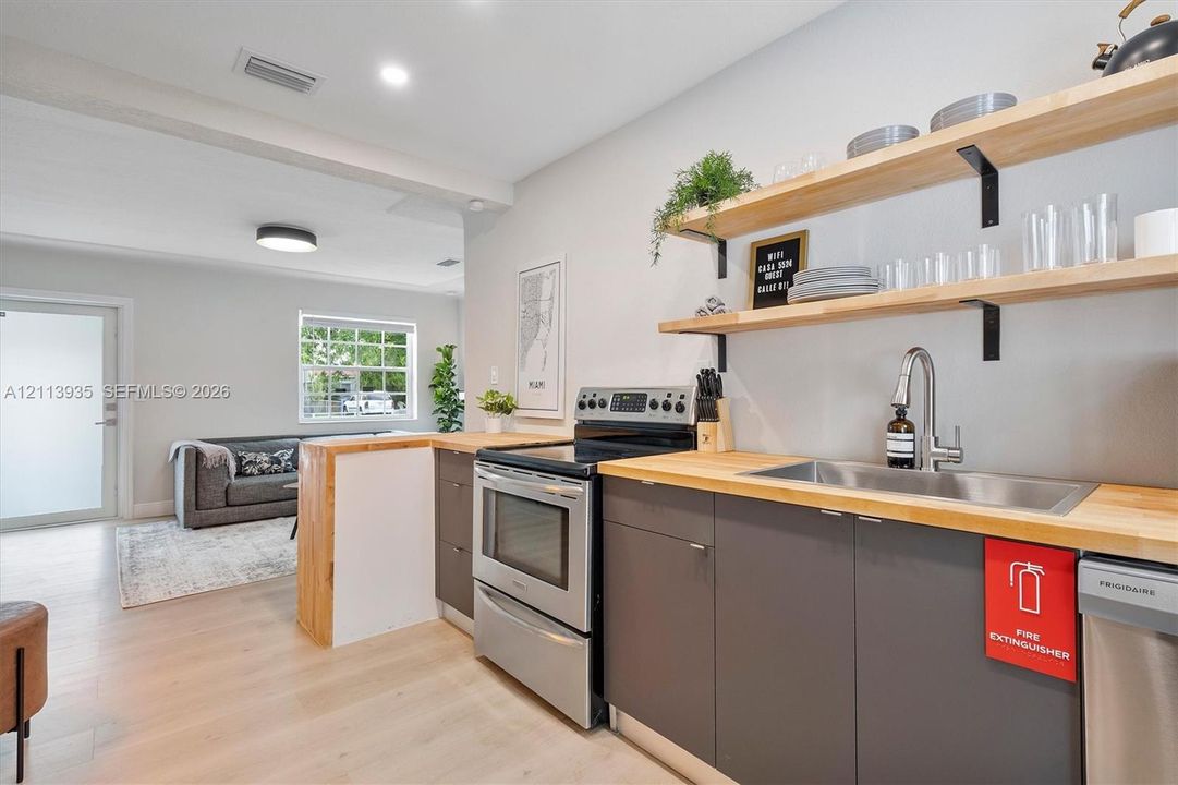 Kitchen area featuring wood-finish countertops, grey cabinetry, stainless steel range and dishwasher, and open wood shelving with metal brackets