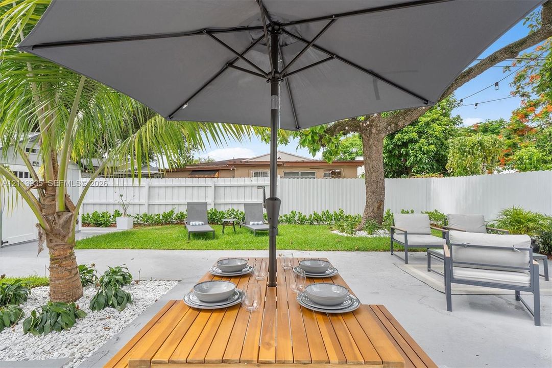Backyard featuring a paved patio, white vinyl fencing, mature trees, and a landscaped garden bed with white stone aggregate