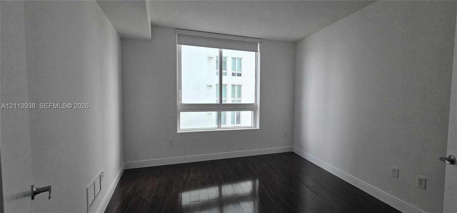 Interior room featuring dark wood-finish flooring, neutral walls, and a large window with a roller shade