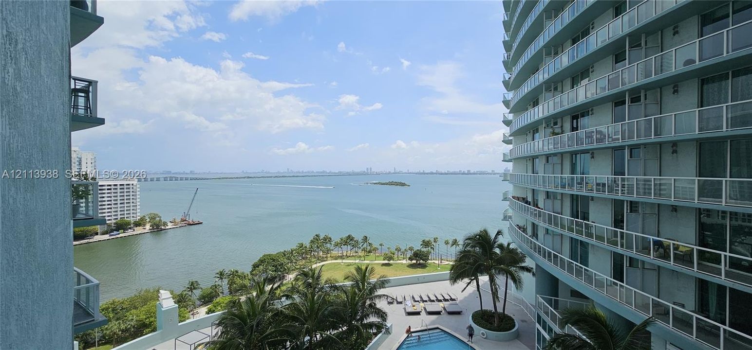 High-rise balcony with glass-panel railings, overlooking a water-facing pool deck, palm trees, and coastal water