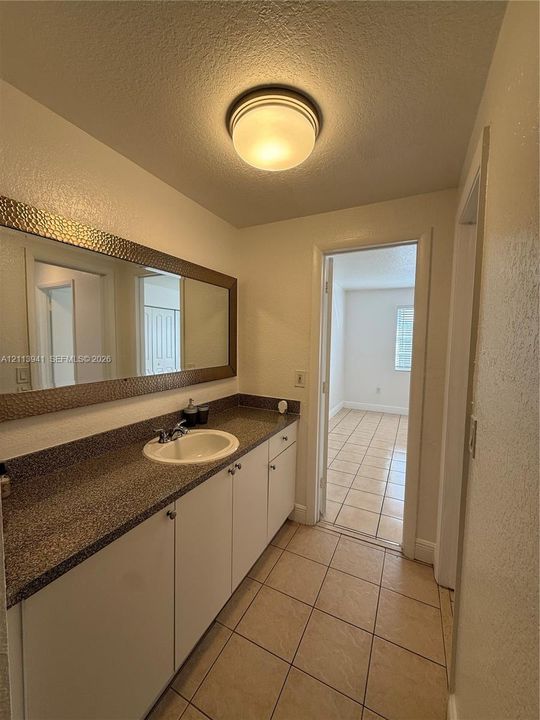 Extended vanity featuring a speckled stone-finish countertop, white cabinetry, a circular undermount sink, and a full-width mirror with a hammered metal-finish frame, complemented by ceramic tile flooring