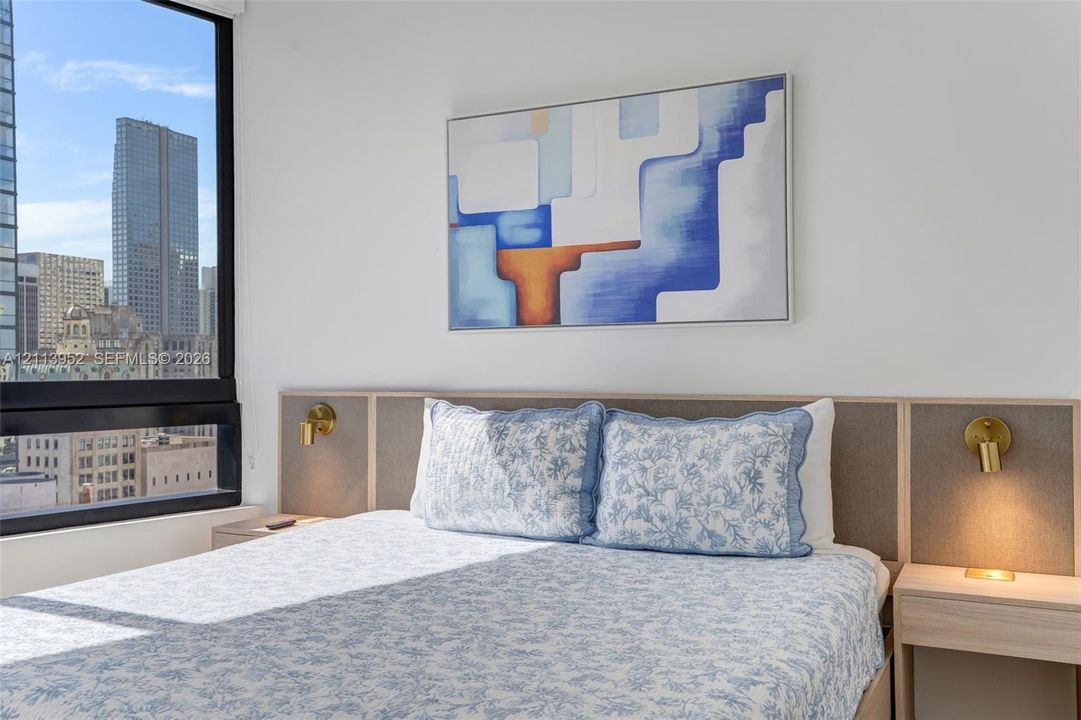 Floor-to-ceiling window, custom wood-paneled headboard with integrated nightstands, and wall-mounted metallic sconces