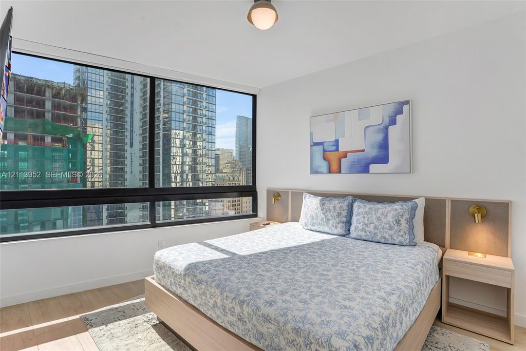 Floor-to-ceiling windows, integrated wood-finish headboard with built-in side tables, recessed ceiling light, and light wood-finish flooring