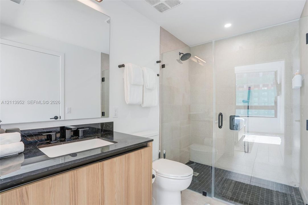 Bathroom featuring a vanity with light wood-finish cabinetry, dark stone countertop, integrated sink, frameless glass-enclosed shower with a rainfall showerhead, and a wall-mounted mirror