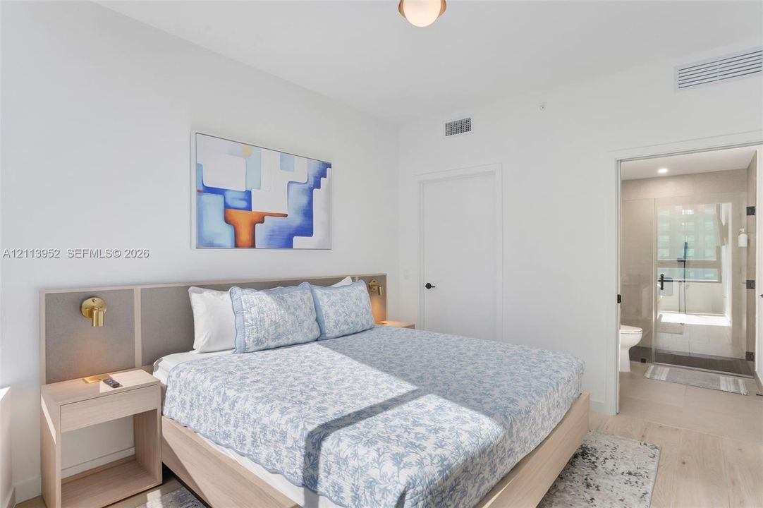 Neutral-toned bedroom featuring a built-in wood-finish headboard with integrated bedside tables, brass wall-mounted light fixtures, light-toned flooring, and a glass-enclosed shower stall