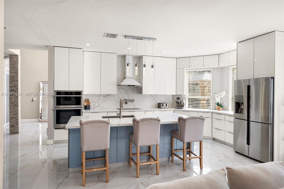 Kitchen featuring white gloss cabinetry, a central island with blue base cabinetry, stone-finish countertops and backsplash, stainless steel built-in double wall ovens, and stainless steel French door refrigerator