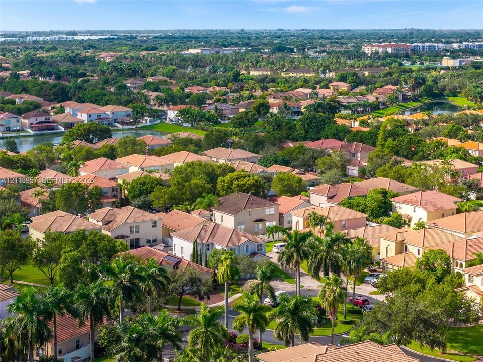 Aerial perspective of a suburban neighborhood featuring paved roadways, multi-story residential structures with tile roofing, mature landscaping, and interspersed bodies of water